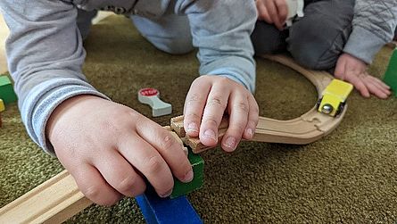 Im Bild sieht man die Hände von zwei Kindern, die Holzeisenbahnschienen zusammenbauen.