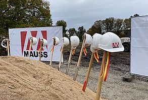 Ein Sandberg mit neun mit gelb-roten Bändern geschmückten Bauspaten. Im Hintergrund sieht man das unbebaute Grundstück und zwei Bauzäune mit dem Logo der Firma MAUSS BAU.