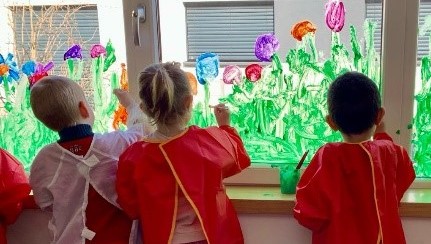 Kinder im Malkitteln bemalen die Fenster mit Fingerfarben.