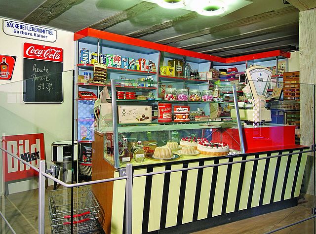 Ein historischer Kaufladen im Stil der 50er Jahre mit Produkten und Marken aus dieser Zeit in den Regalen. Im Vordergrund steht eine gläserne Theke mit einer manuellen Lebensmittelwaage und verschiedene Kuchen, Likörflaschen und Keksdosen.