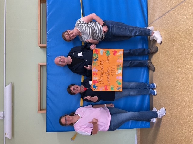 Drei Frauen und ein Mann stehen in einem Turnraum vor einer Weichbodenmatte mit einem Plakat, das den Text "Elterninitiative der Kinderonkologie Erlangen 1100,-" beinhaltet, als Spendenscheck-Symbol. Das Plakat ist mit buten Hndabdrücken an den Rändern verziert.