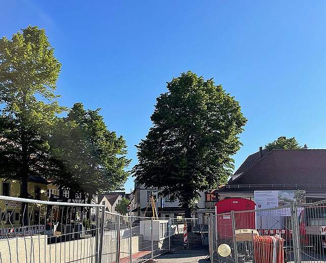 Ein Stadtplatz mit einer Großbaustelle, eingerahmt von Bauzäunen und mit gelagerten Baumaterialien. Im Hintergrund sieht man ein einstöckiges historisches Gebäude und Bäume.