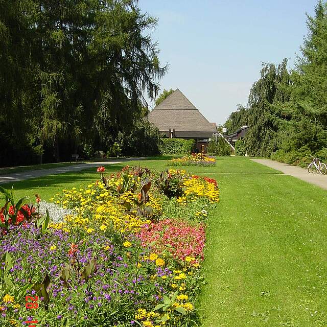 Aussegnungshalle am Neuen Friedhof in Forchheim Im Hintergrund die Aussegnungshalle am Neuen Friedhof in Forchheim. Im Vordergrund bunte Sommerbeete und Wiesenfläche umrahmt von hohen Bäumen.