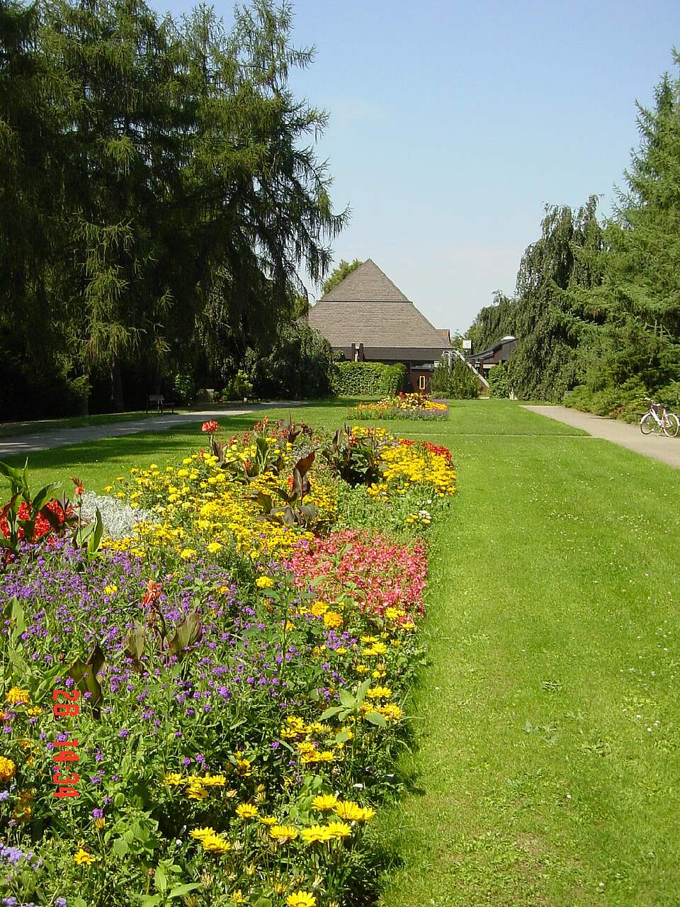 Im Hintergrund die Aussegnungshalle am Neuen Friedhof in Forchheim. Im Vordergrund bunte Sommerbeete und Wiesenfläche umrahmt von hohen Bäumen.