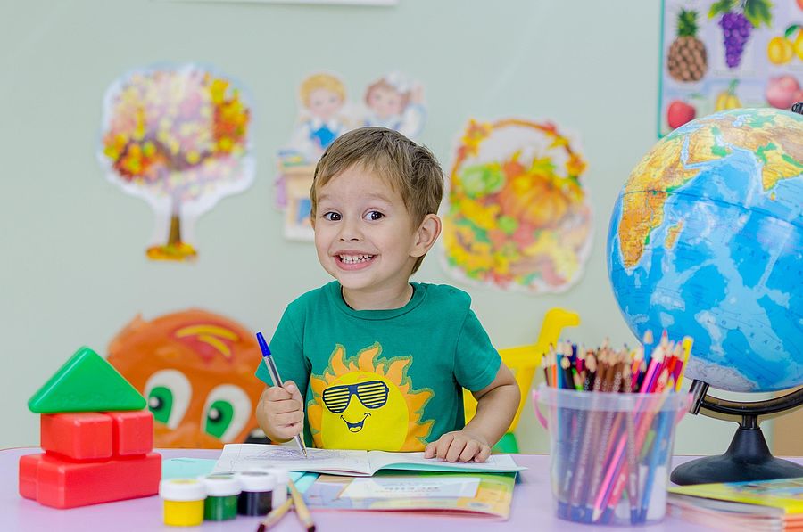 Ein fröhlicher Junge malt an einem Schreibtisch, auf dem bunte Stifte, Bauklötze und ein Globus stehen. Die Wand im Hintergrund ist bunt beklebt.