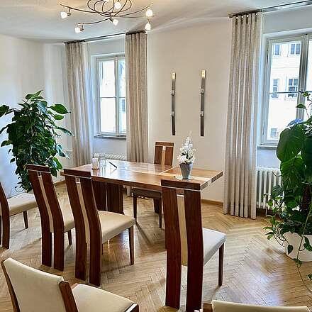 Das Trauungszimmer in Forchheim mit modernen Holzmöbeln, zwei großen Grünpflanzen und einem bunten Wandbild. Auf dem Tisch des Standesbeamten stehen eine weiße Orchidee, eine weiße Kerze und zwei Füller.