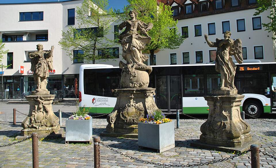 Drei Figuren auf einem Platz, im Hintergrund ist ein Omnibus und eine Häuserfassade zu sehen. 