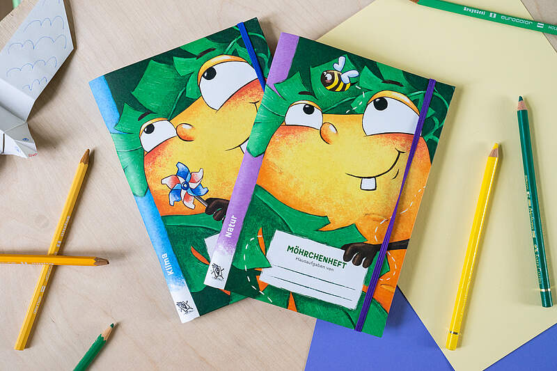 Möhrchenheft für Grundschülerinnen und -schüler Zwei aufgefächerte Hausaufgabenhefte mit dem Möhrchenmotiv, einer Karotte mit Gesicht im Comic-Stil, leigen auf einem Tisch umgeben von Papier und Stiften.