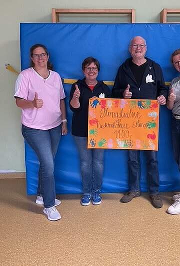 Drei Frauen und ein Mann stehen in einem Turnraum vor einer Weichbodenmatte mit einem Plakat, das den Text "Elterninitiative der Kinderonkologie Erlangen 1100,-" beinhaltet, als Spendenscheck-Symbol. Das Plakat ist mit buten Hndabdrücken an den Rändern verziert.