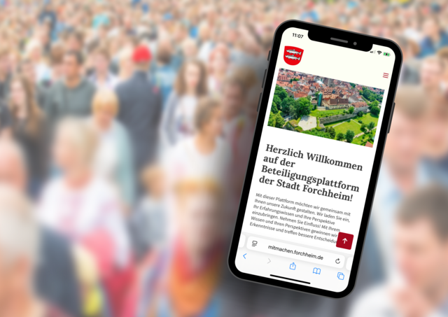 Ein Smartphone-Display zeigt eine digitale Beteiligungsplattform. Im Bildhintergrund erkennt man schehmenhaft eine große Menschenmenge