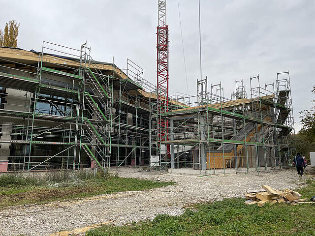 Die eingerüstete Außenfassade der Kita Merowingerstraße Der Rohbau der neuen Kita Merowingerstraße ist umgeben von Baustellengerüsten. In der Mitte steht ein Baukran. Im Vordergrund wechseln sich Wiesen- und Schotterflächen ab. Rechts vorne im Bild liegt ein Stapel mit Holzbaumaterial.
