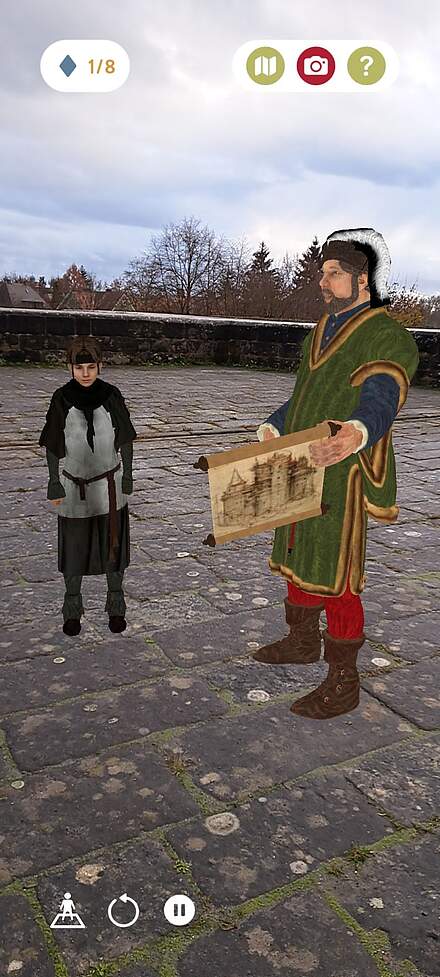 Screnshot aus der App Via Forte Screenshot einer Erlebnis-App, bei der historische virtuelle Personen auf einer Festungsmauer stehen. Ein Mann hält dabei eine Pergamentrolle mit einer Zeichnung in der linken Hand.