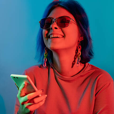 Fröhlich lachende Frau benutzt Smartphone im roten Neonlicht, vor blauem Hintergrund.