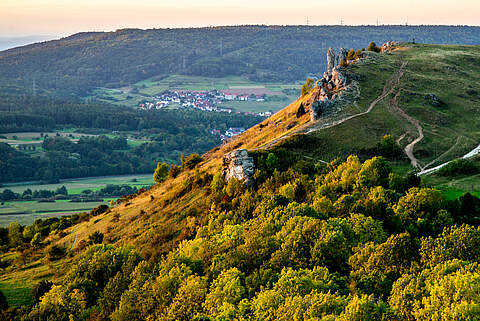 Das "Walberla" bei Sonnenuntergang nahe der Stadt Forchheim. Schrägbildaufnahme von einem Berg bei Sonnenuntergang. Im Hintergrund befindet sich befindet sich ein Dorf, eingerahmt von Wald.