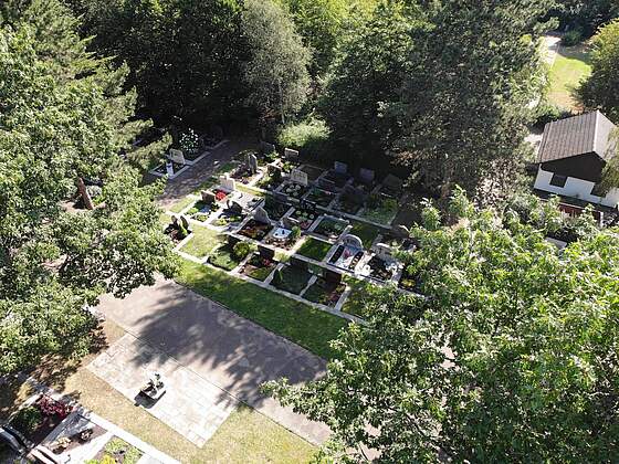 Luftbildaufnahme eines Friedhofs mit bunt bepflanzten Gräbern, umrahmt von großen Bäumen