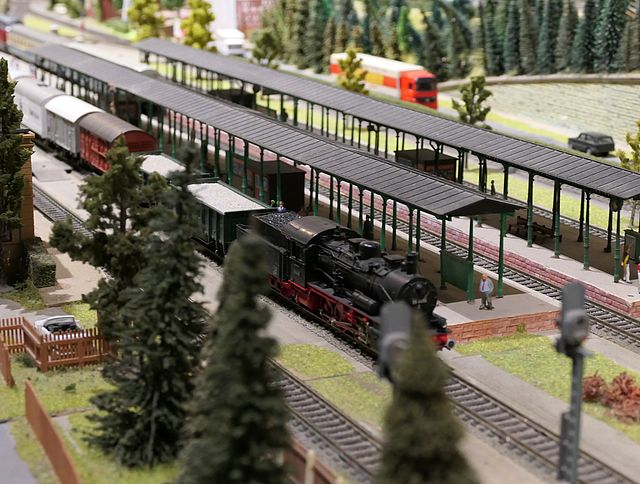 Eine Modelleisenbahn-Landschaft zeigt einen Zug mit einer Dampflok an einem Bahnhof stehen. Im Hintergrund befinden sich auf einer Straße Pkws und zwei Lastwagen.