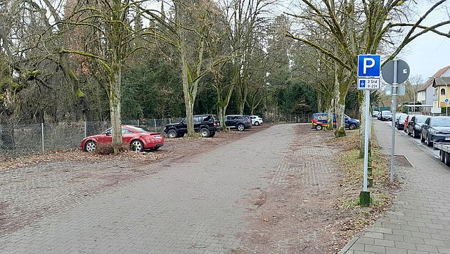 Ein gepflasterter Parkplatz umgeben von Bäumen und Sträuchern. Einige Autos parken darauf. Rechts im Bild befindet sich ein Straßenzug mit Wohnhäusern und parkenden Autos.