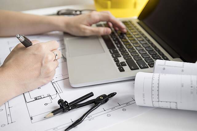 Baupläne und ein Laptop befinden sich auf einem Tisch. Eine Person zeichnet an einem Plan und tippt zeitgleich am Laptop. Im Vordergrund liegt ein Zirkel.