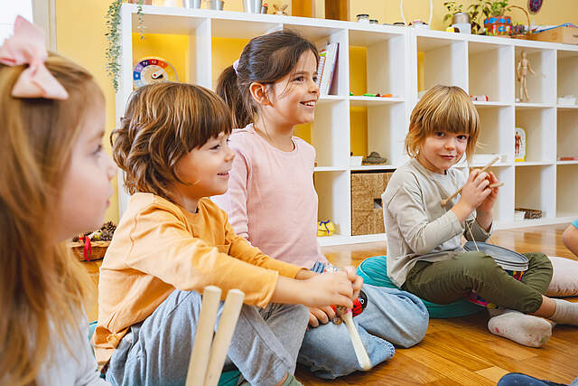 Kindergartenkinder im Morgenkreis in einen Gruppenraum. Im Hintergrund steht ein offenes weißes Regal mit Spielsachen.
