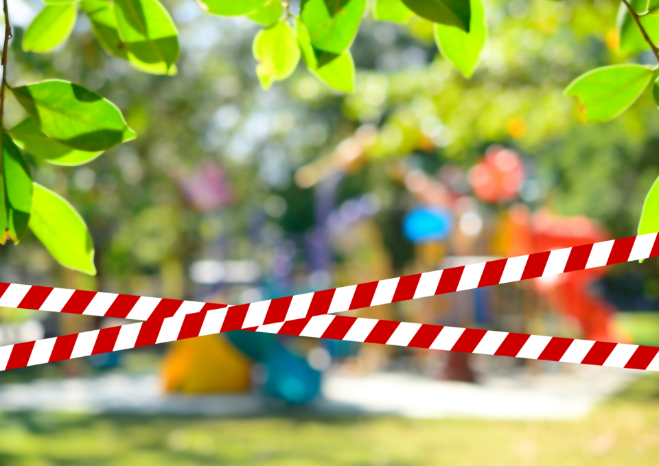 Spielplatz geschlossen Im Hintergrund sieht man schemenhaft einen Spielplatz. Im Vordergrund ist gekreuzt ein Absperrband angelegt und darüber der Text "Spielplatz kurzzeitig geschlossen.