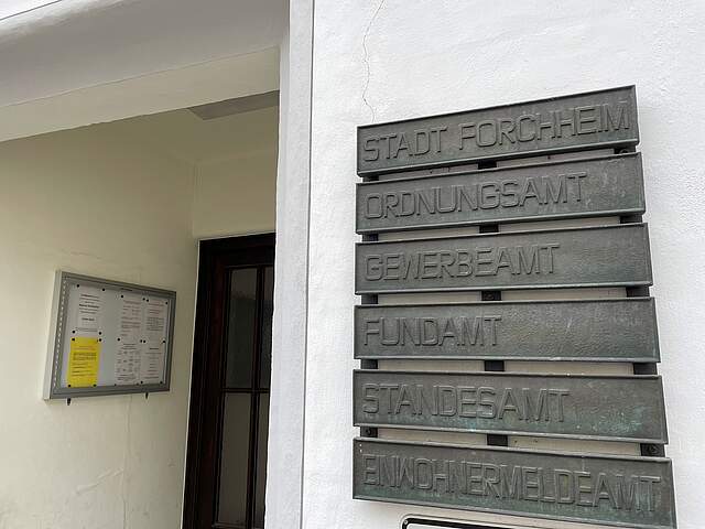 Ein Eingangsbereich einer Behörde mit sechs Metalltafeln an der Außenwand, die die Ämter innerhalb des Gebäudes auflisten.