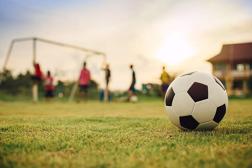 Ein Fußball auf einer Wiese im Vordergrund. Dahinter verschwommen Kinder beim Fußballspielen an einem Tor.