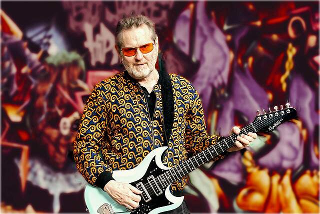 Ein Rockmusiker mit gemustertem Hemd, weißer E-Gitarre und Brille mit orangefarbenen Gläsern vor einem bunt gemusterten Hintergrund.