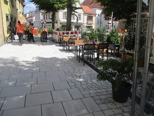 Gepflasterer breiter Fußweg neben Häusern. Im Hintergrund befinden sich Tische und Stühle einer Außengastronomie. Der Weg wird gesäumt von Bäumen.