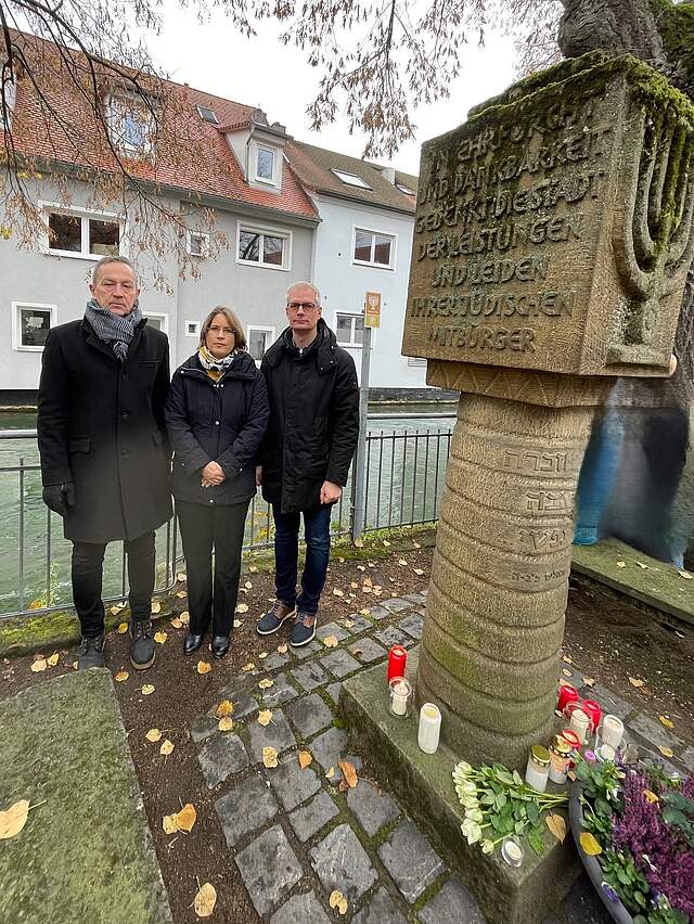 Gedenken an die Opfer der Reichspogromnacht in Forchheim am Von links: Bürgermeister Schönfelder, Bürgermeisterin Dr. Annette Prechtel und Oberbürgermeister Dr. Kirschstein stehen dunkel gekleidet an einem Synagogendenkmal (rechts im Foto). Davor befindet sich ein geschmückter Kranz sowie Grablichter. Im Hintergrund sieht man einen Fluss und eine Häuserzeile.