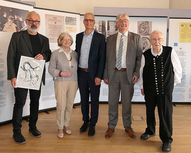 Eine Gruppe aus vier Männern und einer Frau vor mehreren Infowänden mit Fotos, Urkunden und Infotexten. Der Mann links im Bild hält eine Zeichnung in den Händen. Das Motiv zeicgt ein Mädchen mit geflochtenen Zöpfen und ein Haus.