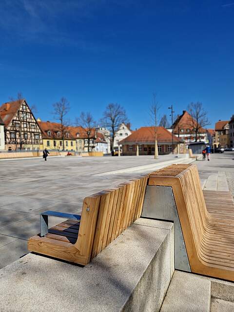 Ein großer, moderner Stadtplatz mit Sitzmöbel aus Holz. Im Hintergrund das historische, einstöckige Sandtsteingebäude "Alte Wache". Die Platz ist von Häusern umgeben.