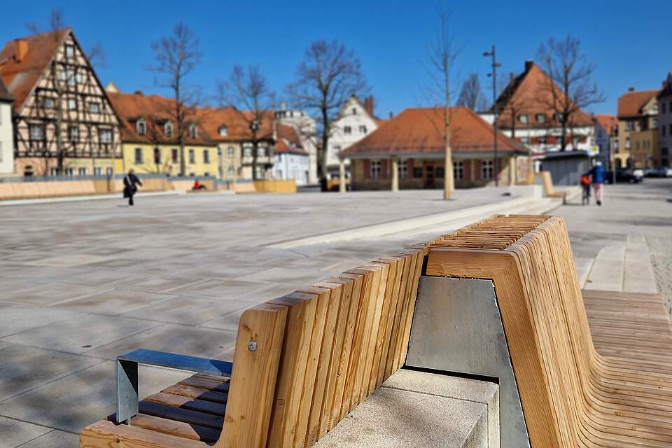 Ein großer, moderner Stadtplatz mit Sitzmöbel aus Holz. Im Hintergrund das historische, einstöckige Sandtsteingebäude "Alte Wache". Die Platz ist von Häusern umgeben.