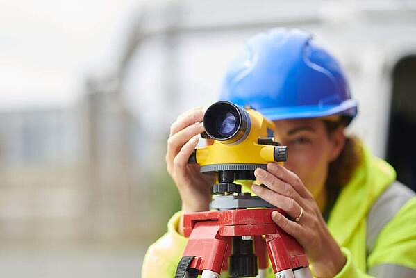 Vermessung Eine Vermessungsingenieurin sieht durch ein Vermessungsgerät. Im Hintergrund ist schemenhaft eine Baustelle zu sehen.