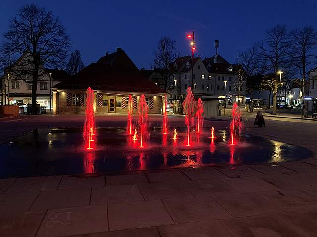 Ein Wasserfontänenfeld bei Nacht mit rot angestrahlten Fontänen auf einem Stadtplatz.