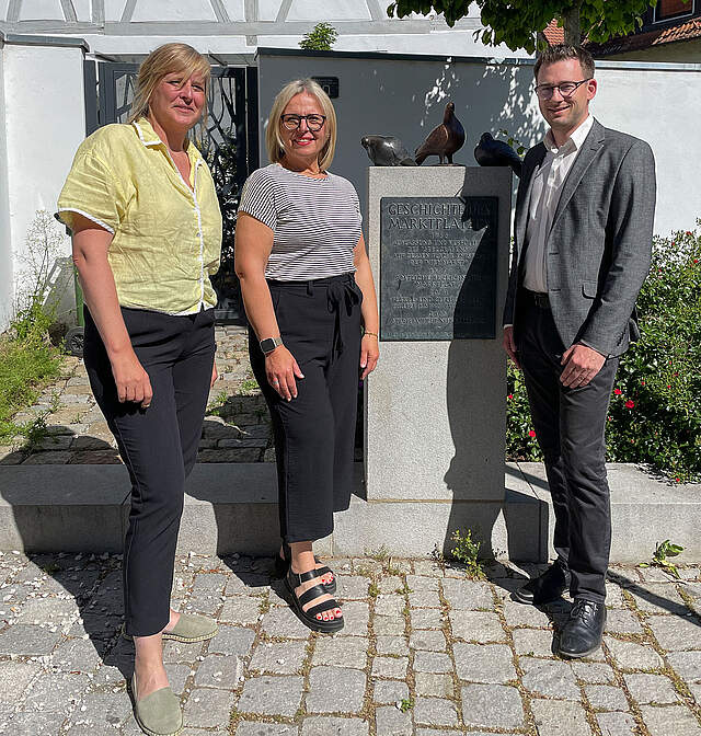 Bronzetauben Schenkung Heimatverein Zwei Frauen und ein Mann umrahmen ein Kleindenkmal mit drei Bronzetauben an einem Stadtplatz.