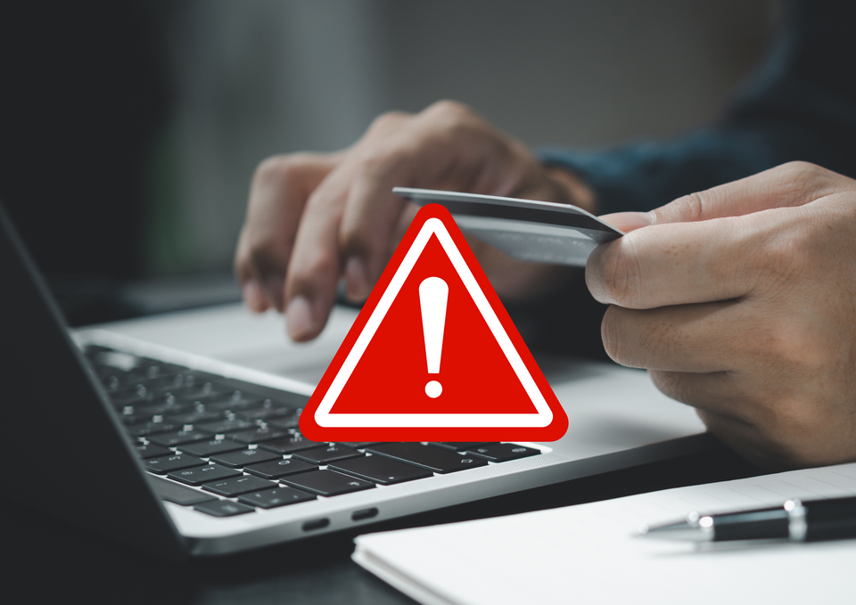 Warnung vor betrügerischen E-Mails Ein aufgeklappter Laptop sowie ein Schreibblock mit einem Kugelschreiber befinden sich auf einem Tisch am dem eine Person mit einer Kreditkarte sitzt und tippt