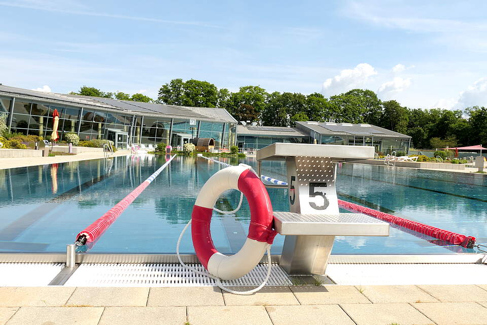 Außensportbecken eines Freizeitbads mit Rettungsreifen an einem Startblock angelegt. Im Hintergrund das Hallenbad mit Saunabereich.