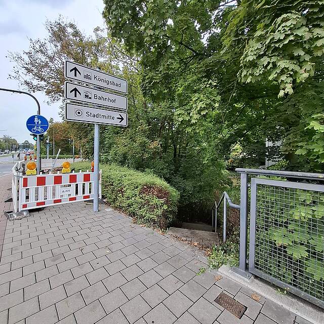 Ein Gehsteig mit einer Barke vor einer Baustelle. Rechts im Bild eine Hecke mit Durchlass zu einem Treppenabgang.