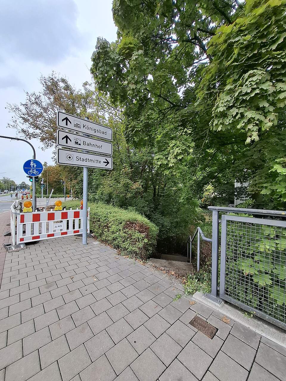 Ein Gehsteig mit einer Barke vor einer Baustelle. Rechts im Bild eine Hecke mit Durchlass zu einem Treppenabgang.