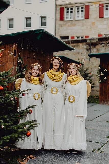 Drei als Engel verkleidete junge Frauen stehen vor festlich geschmückten Weihnachtsbuden.
