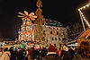 Ein abendlicher Weihnachtsmarkt mit einer beleuchteten Weihnachtspyramide vor einem großen mit weihnachtsmotiven angestrahltem Gebäude.