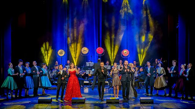 Musikerinnen und Musiker sowie Sängerinnen und Sänger in eleganter Abendgarderobe auf einer blau-gelb erleuchteten Bühne beim Auftritt.