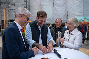 Drei Männer und eine Frau stehen an einem weißen runden Tisch und befestigen gemeinsam den Deckel eine silbrigen Edelstahl-Kapsel.