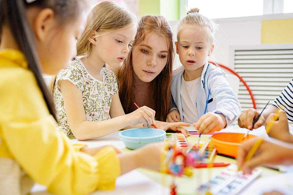 Vorschulkinder mit Erzieherin beim Malen in einem Grußßenraum an einem Tisch.