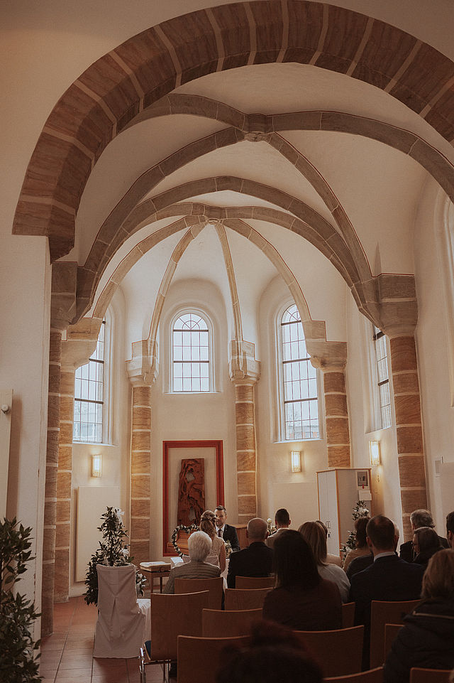 Blick von der Empore hinunter auf eine Hochzeitsgesellschaft in St. Gereon. Zu sehen ist auch das hohe Kreuzgewölbe. Hinter dem Standesbeamten hängt ein ziegelrotes Kunstwerk.
