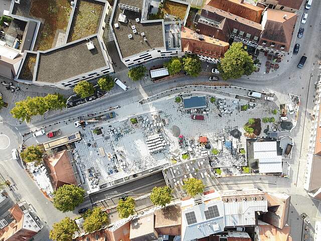 Eine Luftbildaufnahme zeigt die Umbaumaßnahmen auf einem großen Stadtplatz mit Pflasterarbeiten, Baumaterial und neuen Bäumen.
