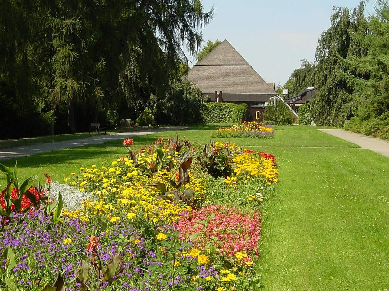 Aussegnungshalle im Neuen Friedhof aus den 80er Jahren. Im Vordergrund befindet sich eine große Rasenfläche mit einer sommerlich bepflanzten Staudenrabatte.