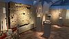 Ein Ausstellungsraum in einem Museum zeigt ein Sedimentspräparat, das an der Wand hängt umgeben von Glasvitrinen. Im linken Bildrand ist ein Absperrband erkennbar mit dem Schriftzügen "Einsatz" und "Archäologie".