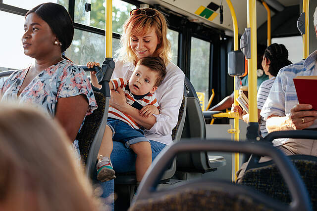 Mehrere Personen sitzen in einem Bus. Eine Frau hält ein Kleinkind auf ihrem Schoß.