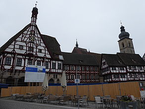 Ein langer Holzbauzaun umgibt ein großes historisches Fachwerkgebäude. Innerhalb der Absperrung stehen blaue Baucontainer, ein Gerüst und eine große Bautafel vor dem Gebäude.